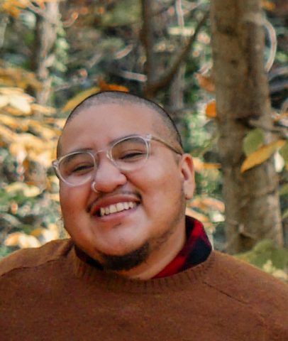 Ollin, wearing a brown sweater over red and black flannel, smiles at the camera. They wear clear-framed glasses. Autumnal trees are blurred in the background.